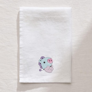 May include: White rectangular towel with an embroidered cartoon character in light blue, purple, and pink. The character has a light blue head, purple hair, a pink nose, and is sitting with its hands clasped.