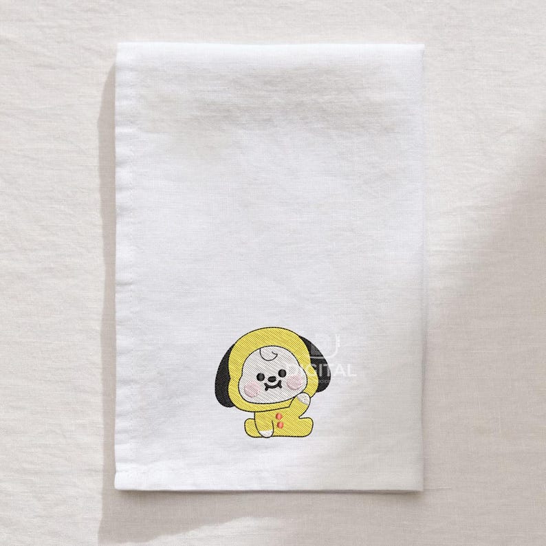 May include: White rectangular towel with an embroidered yellow cartoon character wearing a hooded outfit. The character has black ears and a smiling face. The towel is folded and appears to be made of a soft fabric.