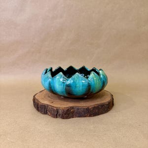 May include: A teal and black ceramic bowl with a scalloped rim, resembling a flower, sits on a round wooden slice. The bowl's interior is dark, contrasting with the vibrant teal exterior. The wooden base adds a natural element to the decorative piece.