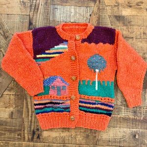May include: An orange knitted cardigan with a landscape design. The sweater features a house, tree, and colourful stripes. It has a round neck, long sleeves, and wooden buttons. The sweater is made of a thick, textured yarn.