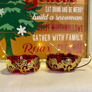 May include: Two red glass candle holders with gold floral filigree. One is a cup shape with a handle. The other is a round bowl shape. The background is a wooden sign with holiday phrases.