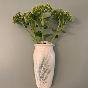 May include: A cream ceramic wall vase with green and blue splatter design, holding green flowering stems. The vase has a rounded body and a narrow neck, mounted on a grey wall.