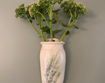 May include: A cream ceramic wall vase with green and blue splatter design, holding green flowering stems. The vase has a rounded body and a narrow neck, mounted on a grey wall.