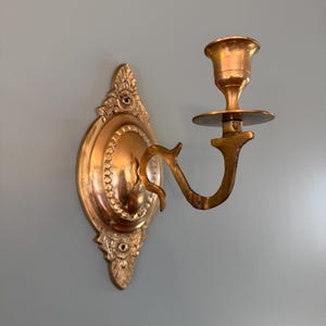 Peut inclure: Applique murale dorée ornée avec un bougeoir. La plaque arrière décorative présente des détails floraux et des volutes, et un bras incurvé supporte la coupelle à bougie. Le métal a une finition chaude et vieillie.