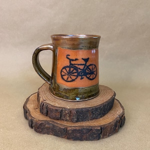 May include: A ceramic mug with a bicycle design. The mug has a brown handle and rim, with an orange centre panel featuring a black bicycle illustration. The mug sits on two wooden coasters with a natural bark edge.