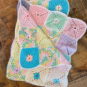 May include: A pastel-coloured, handmade crochet blanket. The blanket features a floral pattern with square motifs and a scalloped edge. The colours include pink, blue, yellow, and green. The blanket is displayed on a wooden surface.