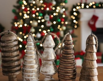 May include: Five wooden Christmas tree decorations with twine hangers. The ornaments are various shapes and shades of brown and white. A decorated Christmas tree and fireplace with stockings are in the background.