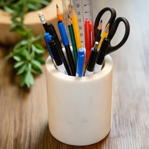 May include: A white cylindrical pencil holder filled with various writing utensils. The holder contains black pens, pencils, a ruler, scissors, and a red pen. The items are arranged in a neat and organized manner, ready for use.