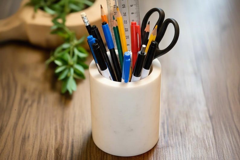 May include: A white cylindrical pencil holder filled with various writing utensils. The holder contains black pens, pencils, a ruler, scissors, and a red pen. The items are arranged in a neat and organized manner, ready for use.