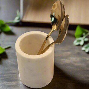 May include: A white marble utensil holder with two gold spoons inside. The cylindrical holder sits on a dark wooden surface, with green foliage in the background. The spoons are made of a shiny gold metal.