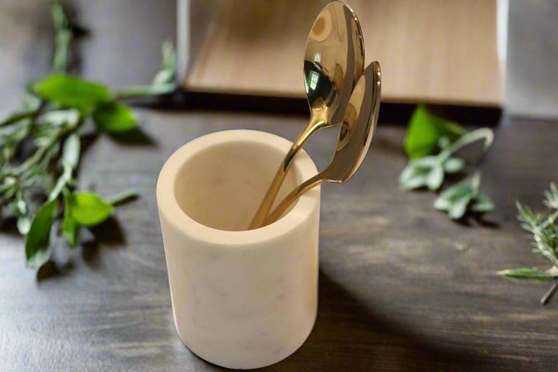 May include: A white marble utensil holder with two gold spoons inside. The cylindrical holder sits on a dark wooden surface, with green foliage in the background. The spoons are made of a shiny gold metal.