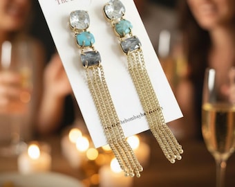 May include: Dangle earrings with a gold-tone metal setting. Each earring features clear, turquoise, and grey gemstones, with multiple strands of gold chains cascading down. The earrings are displayed on a white card.