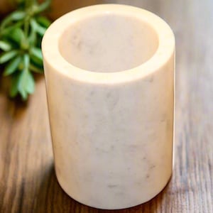 May include: A cylindrical, off-white marble container with a hollowed-out interior. The smooth, polished surface has subtle gray veining. The container is set on a wooden surface, with green foliage visible in the background. The object is approximately 4 inches tall.