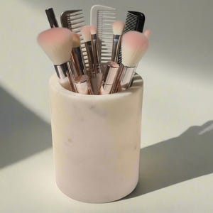 May include: A collection of makeup brushes and combs in a cylindrical marble container. The brushes have pink bristles and silver handles. The container is a light pink and cream color. The image is lit with natural light.