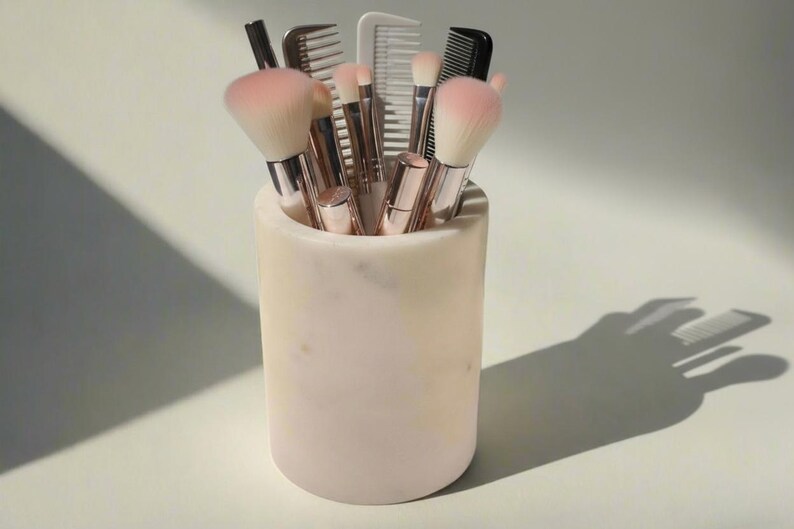 May include: A collection of makeup brushes and combs in a cylindrical marble container. The brushes have pink bristles and silver handles. The container is a light pink and cream color. The image is lit with natural light.