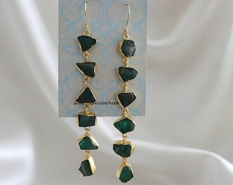 May include: Gold-plated drop earrings with dark green, irregularly shaped gemstones. Each earring has five gemstones, connected by gold links. The earrings hang from a gold hook. The background is a light blue card with text and white fabric.