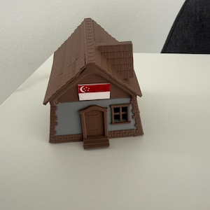 Peut inclure: Une maquette de maison miniature avec un toit marron et des murs peints en gris et marron. La maison comprend une petite porte, une fenêtre et un drapeau de Singapour. Le modèle est un objet décoratif.