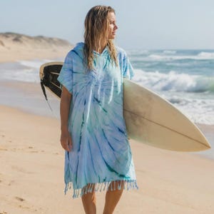 Könnte beinhalten: Ein blau-türkisfarbener Kapuzenponcho mit Fransenbesatz. Der Poncho wird an einem Strand getragen und mit einem Surfbrett gehalten. Das Surfbrett ist cremefarben mit schwarzen Akzenten. Der Ozean und der Sand sind im Hintergrund.