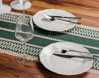 May include: A dining table setting with white plates, silverware, and a wine glass. The plates have a scalloped edge. A green and white patterned table runner is in the center. The table is made of wood.