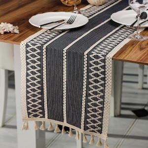 May include: A navy and cream table runner with a geometric pattern. The runner has vertical stripes and a decorative border with tassels. It's on a wooden table, with white plates, cutlery, and wine glasses. The runner's length is approximately 150 cm.