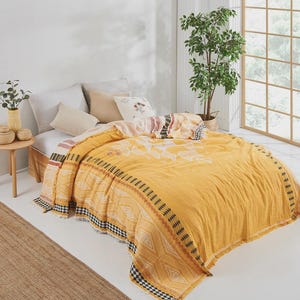 Puede incluir: Una colcha amarilla con un patrón geométrico y bordes con flecos. La colcha presenta una mezcla de diseños de diamantes y zigzag en negro, blanco y naranja. La cama está hecha con almohadas blancas y una almohada beige. La cama está en una habitación luminosa con una ventana.