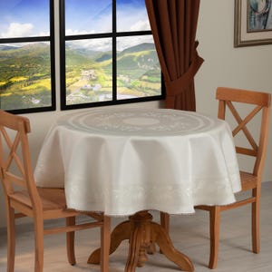 Op de afbeelding: Een ronde, crèmekleurige tafelkleed met een bloemmotief bedekt een houten tafel. Het tafelkleed heeft een decoratieve rand en staat in een kamer met houten stoelen en een schilderachtig uitzicht door het raam.