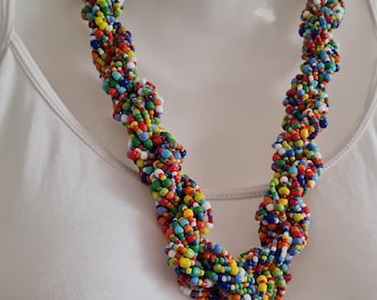 May include: A colorful beaded necklace with a twisted design. The necklace features a variety of small beads in red, yellow, green, blue, and white. The necklace is displayed on a white mannequin.