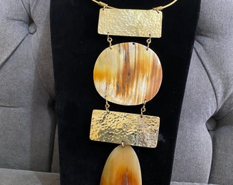Peut inclure: Un collier de déclaration avec un collier et un pendentif en métal doré. Le pendentif comprend un rectangle doré martelé, un pendentif rond aux tons marron et crème, un autre rectangle doré martelé et un pendentif ovale aux tons marron et crème.