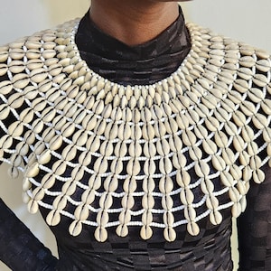 Puede incluir: Un collar de conchas de color crema. El collar está hecho de pequeñas conchas de color claro, intrincadamente tejidas con hilo blanco. Las conchas están dispuestas en un patrón en capas, creando un diseño tipo babero. El collar se usa sobre una blusa negra.