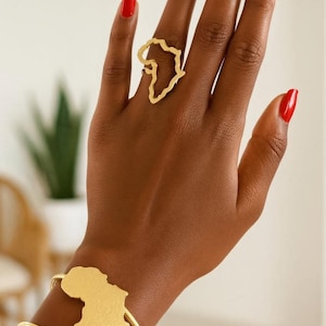 Puede incluir: Conjunto de anillo y pulsera dorados con el contorno del continente africano. El anillo se lleva en un dedo y la pulsera en la muñeca. El fondo es neutro, con una planta visible.