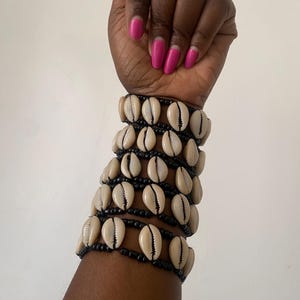 Puede incluir: Varios brazaletes hechos con conchas de cauri de color crema y cuentas negras. Los brazaletes están apilados uno encima del otro. La piel de la persona es oscura y sus uñas están pintadas de color rosa brillante.