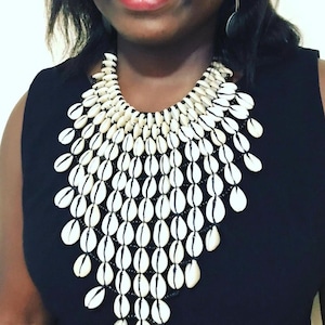 Könnte beinhalten: Eine auffällige Halskette mit zahlreichen weißen Muscheln mit schwarzen Akzenten, die in einem Kaskadenmuster angeordnet sind. Die Halskette ist vor einem schwarzen Hintergrund platziert, was einen starken Kontrast erzeugt. Die Muscheln sind mit schwarzen Perlen aufgefädelt.