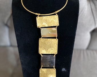 Peut inclure: Un collier doré avec une série de pendentifs rectangulaires. Les pendentifs alternent entre du métal doré martelé et de la pierre naturelle dans des tons crème, noir et orange. Le collier est présenté sur un mannequin noir.
