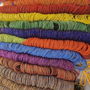 Pode incluir: Uma exibição vibrante de pulseiras de contas coloridas em várias tonalidades, incluindo vermelho, laranja, amarelo, verde, azul e roxo. As pulseiras são empilhadas em fileiras, mostrando uma gama diversificada de cores e texturas.