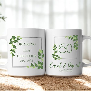 May include: Two white ceramic mugs with green text and leaf designs. One mug reads "DRINKING TEA TOGETHER since 1965". The other mug displays "60" with "Carol & David 12.09.2025".
