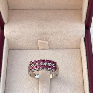 Puede incluir: Un anillo plateado con dos filas de piedras preciosas redondas de color rojo intenso. El anillo se muestra dentro de un joyero beige claro con ribete burdeos. La parte superior de la caja tiene el texto dorado: "KAY ADAMS GEMS & JEWELRY".