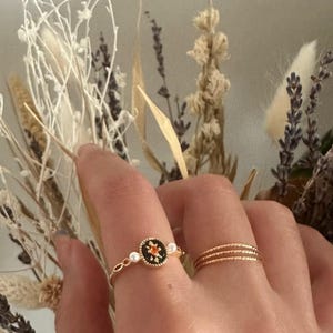 Peut inclure: Une main portant deux bagues en or. L'une présente un motif circulaire noir avec un motif floral orange et deux petites perles. L'autre est une simple bande dorée avec trois anneaux texturés. Des fleurs séchées sont en arrière-plan.