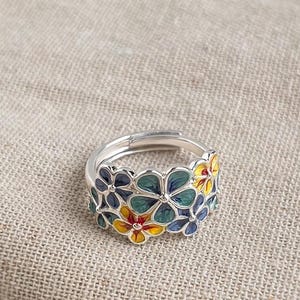 Könnte beinhalten: Ein silberfarbener Ring mit einem Band, das mit mehreren kleinen, fünfblättrigen Blumen verziert ist. Die Blumen sind mit Emaille in den Farben Dunkelblau, Petrol und Gelb ausgefüllt, wobei die gelben Blumen eine rote Mitte haben.