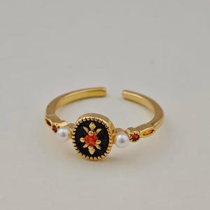 May include: Gold-toned adjustable ring featuring a central black oval with a red floral design. The ring is accented with small pearls and red gemstones. The ring is on a white background.