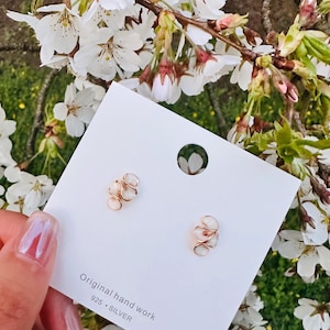 May include: A pair of small, gold-colored earrings with a swirling design, each featuring a small, white, oval-shaped stone. The earrings are displayed on a white card with the text "Original hand work 925 &bull; SILVER". The background includes white flowers.