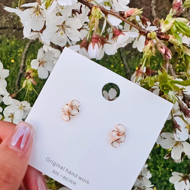 May include: A pair of small, gold-colored earrings with a swirling design, each featuring a small, white, oval-shaped stone. The earrings are displayed on a white card with the text "Original hand work 925 &bull; SILVER". The background includes white flowers.
