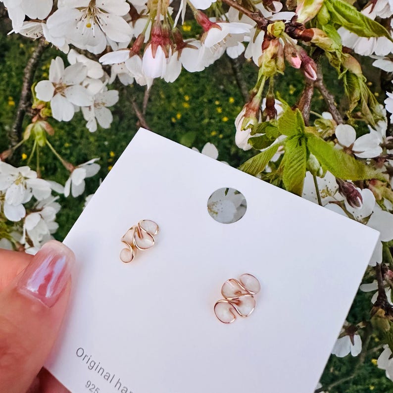 May include: A pair of gold-colored earrings with a unique, swirling design. The earrings are displayed on a white card with the text "Original hand" and "925". The background features white flowers and green foliage.