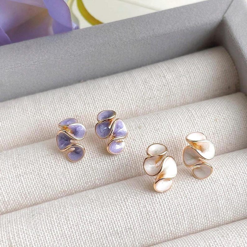 May include: Two pairs of stud earrings. One pair features a gold-tone metal with lavender enamel, and the other pair has a gold-tone metal with white enamel. The earrings are displayed in a gray jewelry box with a white fabric lining.