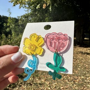 Könnte beinhalten: Ein Paar bunte Acryl-Ohrringe. Ein Ohrring zeigt eine gelbe Blume mit einer blauen Spirale, der andere eine rosa Blume mit einem grünen Blatt. Die Ohrringe sind an einer weißen Karte befestigt.