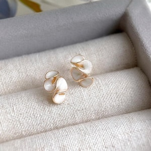 May include: A pair of gold-toned earrings with a white enamel floral design. The earrings have a layered, petal-like appearance, and are set against a neutral, textured background.