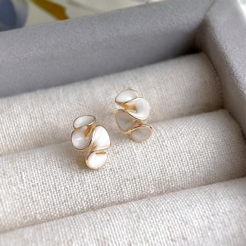 May include: A pair of gold-toned earrings with a white enamel floral design. The earrings have a layered, petal-like appearance, and are set against a neutral, textured background.