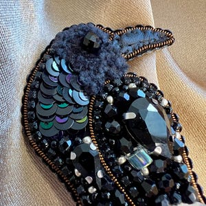 May include: A detailed raven brooch, crafted with black sequins, beads, and crystals. The bird's head is a textured dark blue, and the design is outlined with a brown trim. The brooch is a striking piece of jewellery, perfect for adding a touch of elegance.