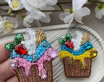 Puede incluir: Dos broches de cupcake con cuentas. Uno es rosa, azul y blanco con una gema roja y gemas rosas. El otro es azul, amarillo y blanco con una gema roja y gemas amarillas. Ambos tienen bases de cuentas marrones.