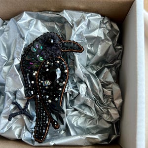 May include: A detailed black bird brooch, crafted with beads, sequins, and crystals, presented in a cardboard box with silver tissue paper. The brooch showcases a textured design with a variety of embellishments, creating a striking visual.