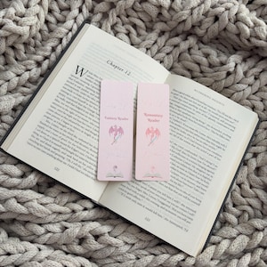 May include: Two rectangular light pink bookmarks resting on an open book. Each bookmark features the text Fantasy Reader or Romantasy Reader, a pink dragon illustration, a sword, and an open book icon at the bottom with a small pink dragon.
